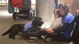Corso sentada leyéndole a la niña que se encuentra a su derecha un libro y la perra de terapia Susana tumbada delante de Corso mirándola expectante